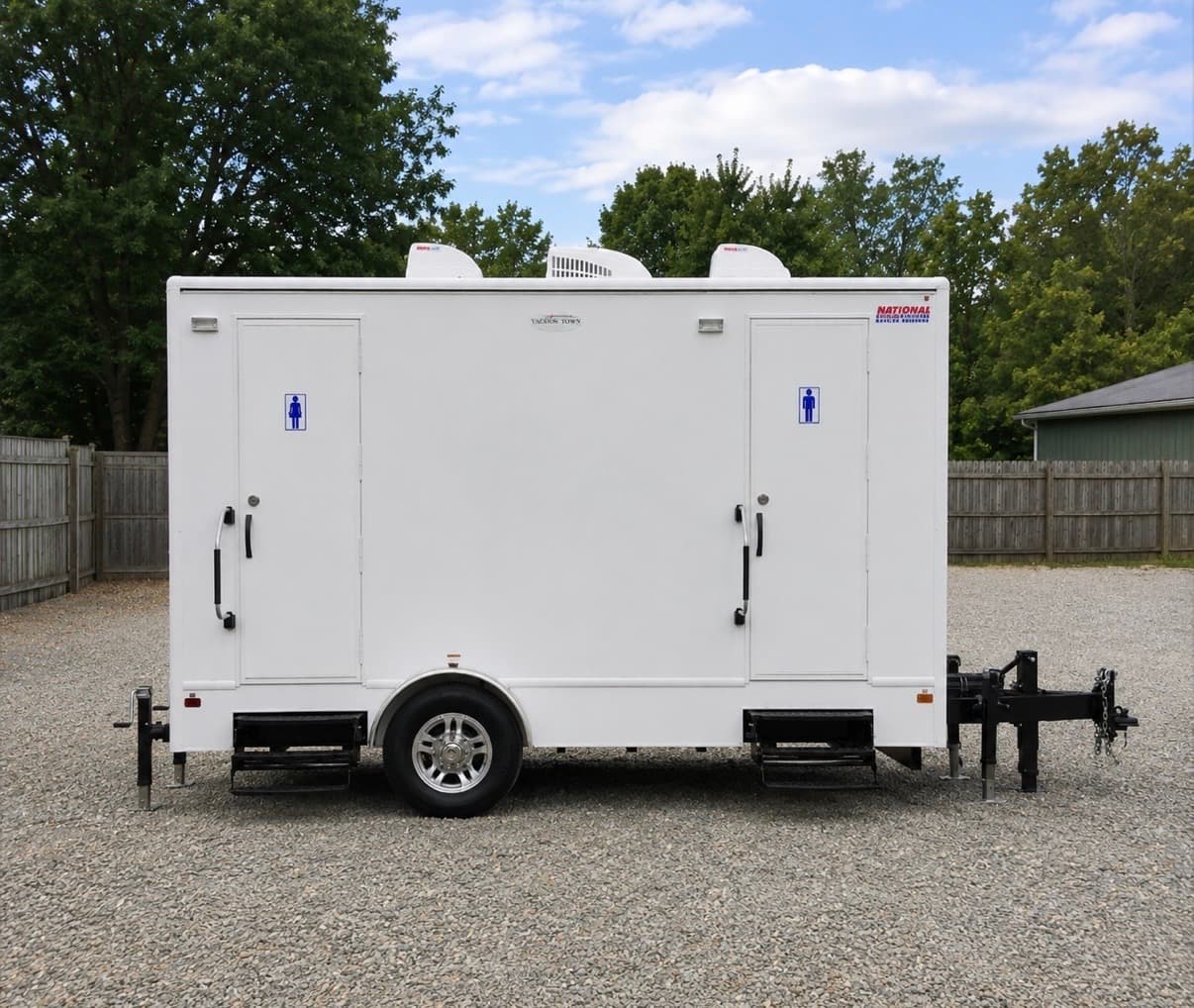 White portable restroom trailer with two doors and steps parked on a gravel lot.