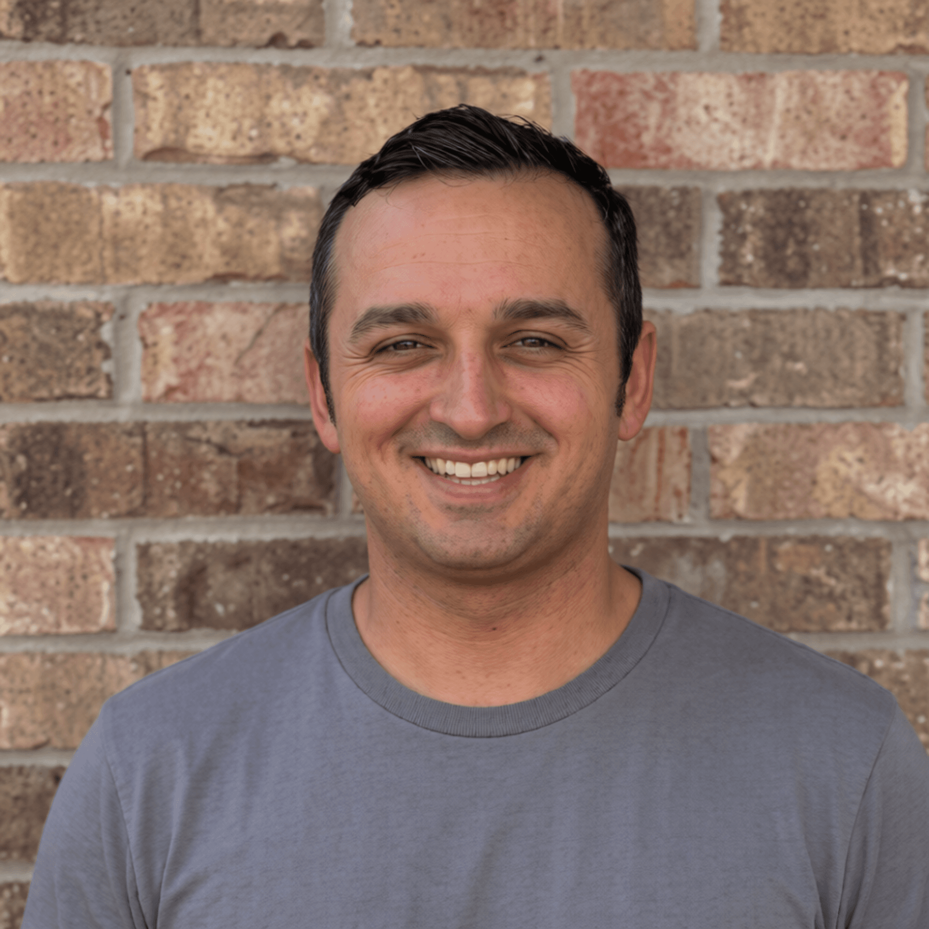 Smiling man with short dark hair wearing a grey t-shirt against a brick wall.
